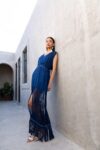 Model wearing the Caribe Blue Maxi Dress, featuring handwoven fringe at the hem and shoulders, styled with white artisan earrings and sandals