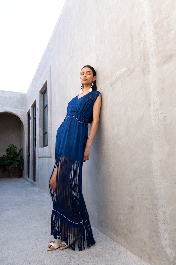 Model wearing the Caribe Blue Maxi Dress, featuring handwoven fringe at the hem and shoulders, styled with white artisan earrings and sandals