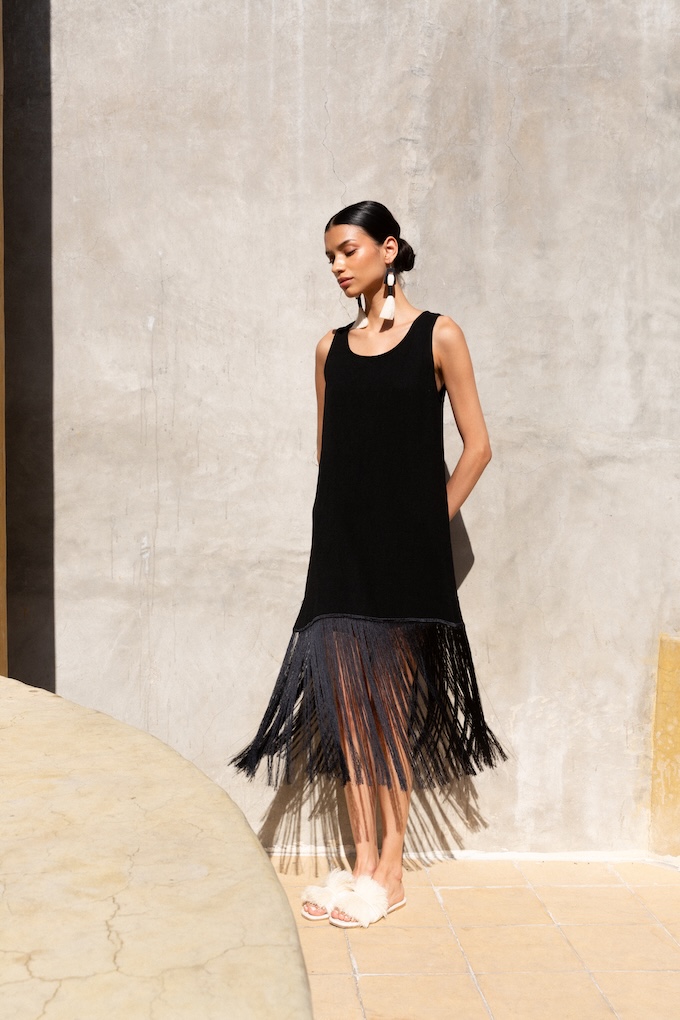 Model wearing Naay Earrings with bull horn and natural fiber fringe, styled with a black dress against a sunlit wall.