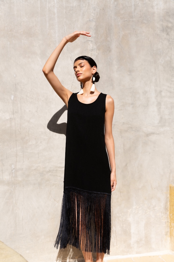Model wearing the Ceiba fringe dress in black, featuring a minimalist silhouette with long flowing fringe. Styled with Maya earrings under natural sunlight