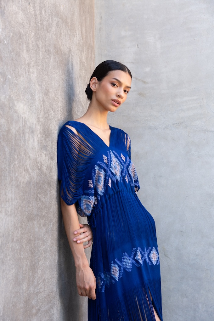 Model wearing the Holbox Blue Maxi Dress with handwoven brocade detail and drawn thread technique, leaning against a textured wall.