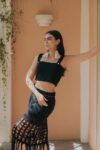 Woman wearing the Tequila crochet crop top in black with matching skirt. Features wide straps and gold button accents, handwoven in Jalisco.