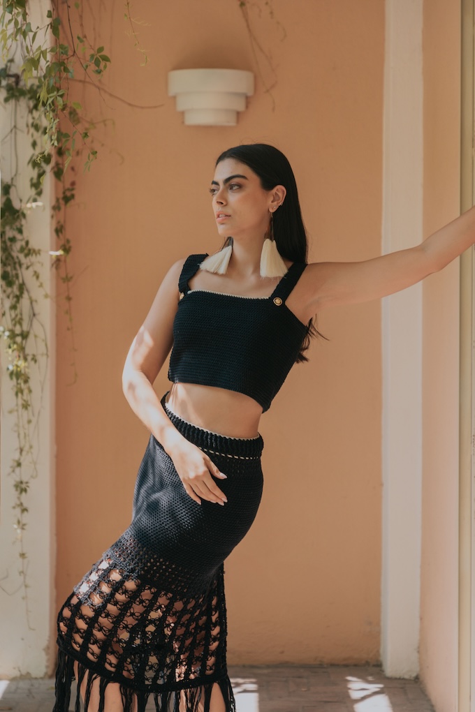 Woman wearing the Tequila crochet crop top in black with matching skirt. Features wide straps and gold button accents, handwoven in Jalisco.