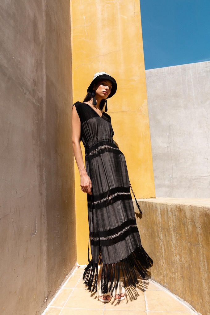 Model wearing the Mezcal Maxi Dress with handwoven fringe and drawn thread detailing, styled with a wide-brim hat in bright architectural setting.